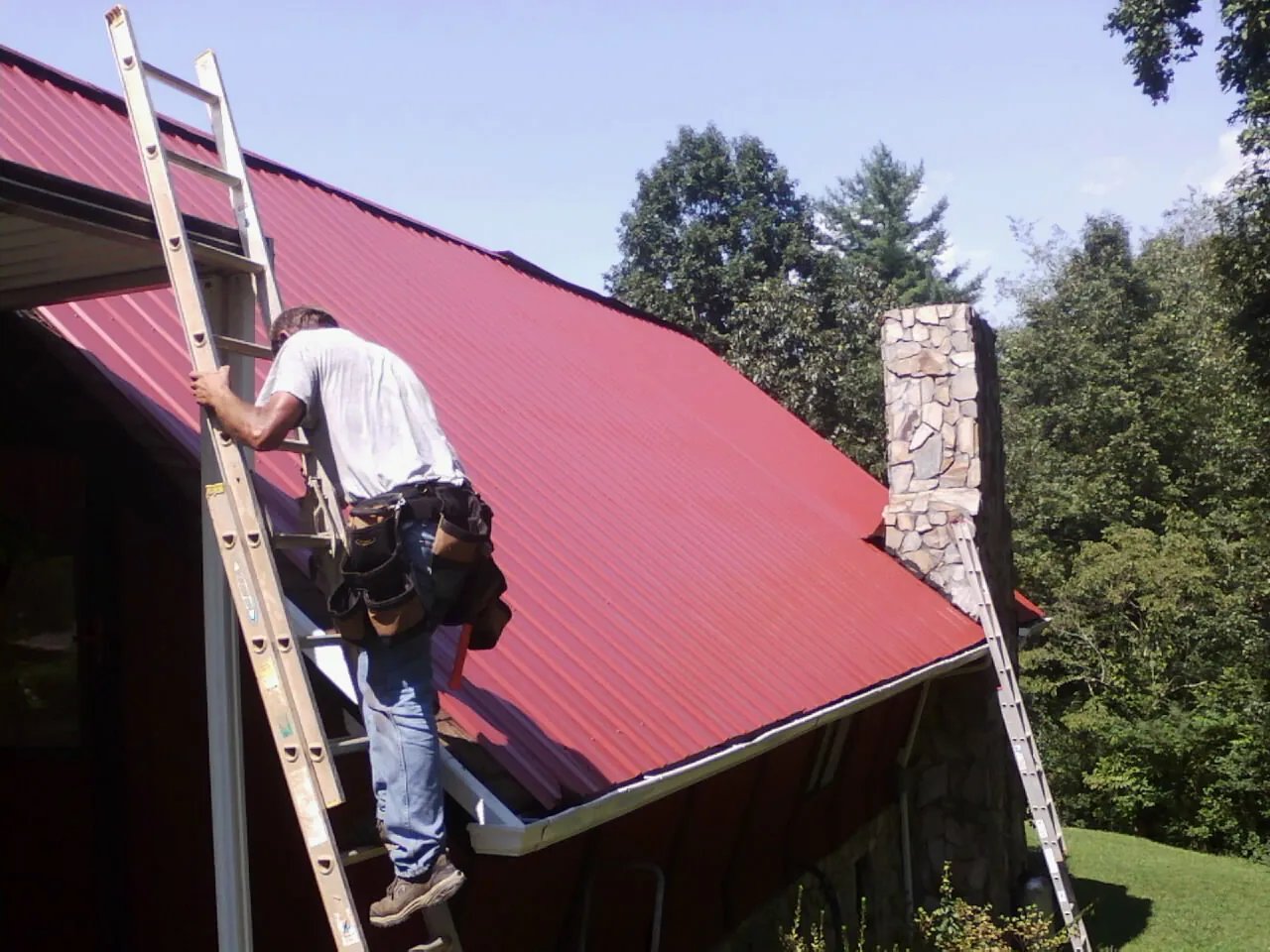 Roofer on a ladder beside a red metal roof and stone chimney — Soffit & Fascia Repair by Roofing Brothers in Eggleston